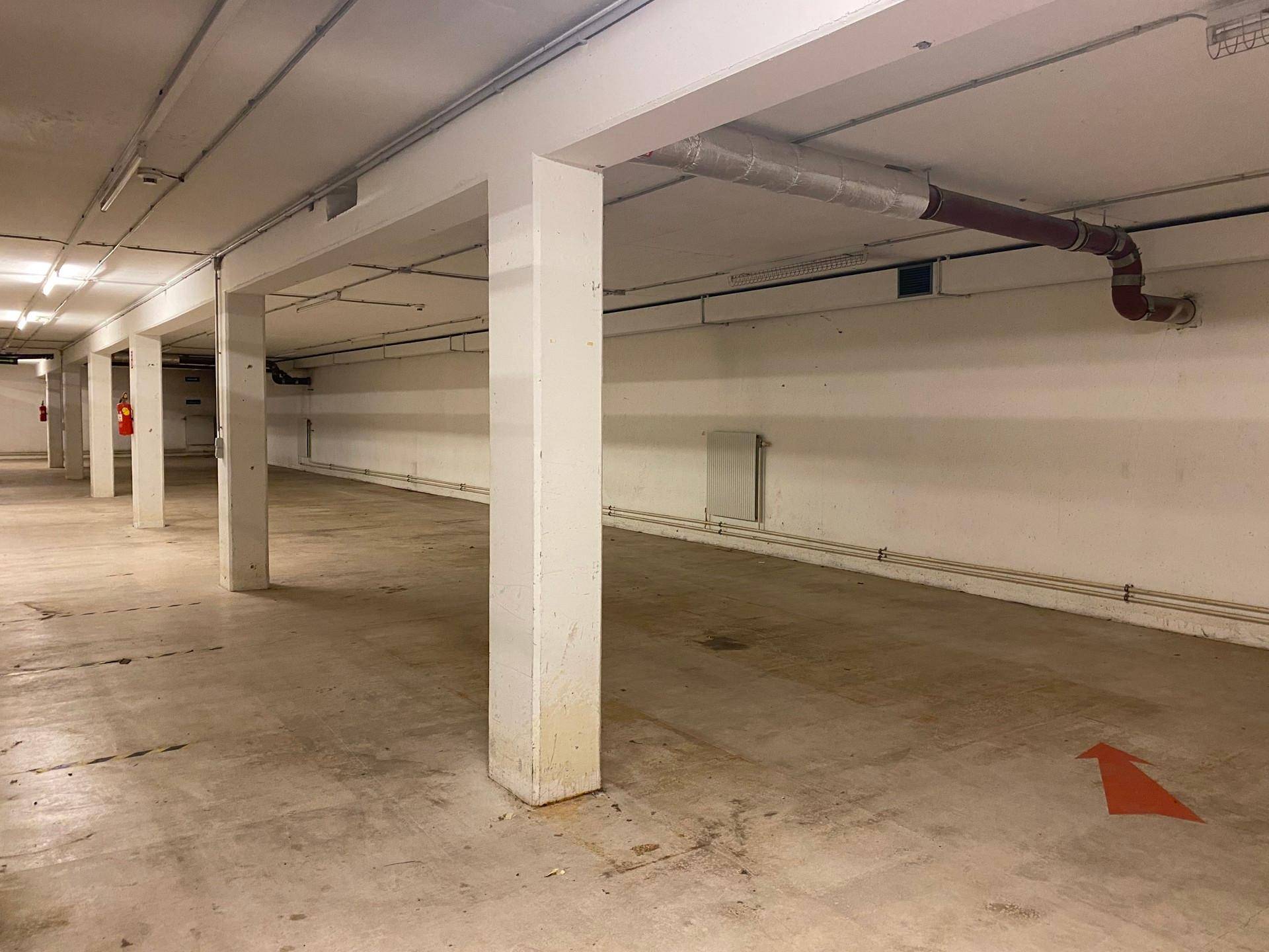 Geräumige Tiefgarage mit Betonstützen und sichtbaren Rohrleitungen an der Decke, gut beleuchtet.