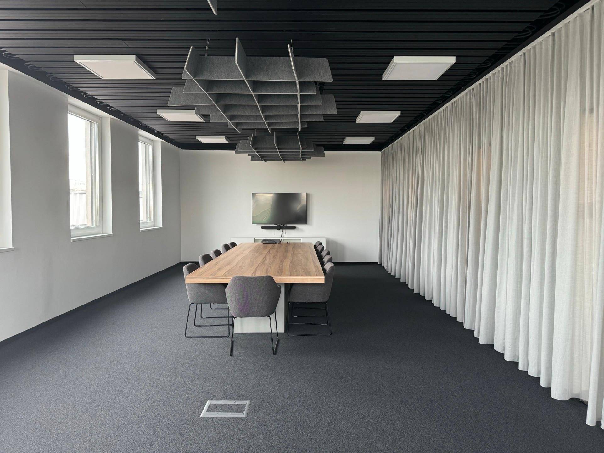 Geräumiger Konferenzraum mit langem Holztisch, bequemen Stühlen, großem Bildschirm und schallabsorbierenden Deckenpaneelen.
