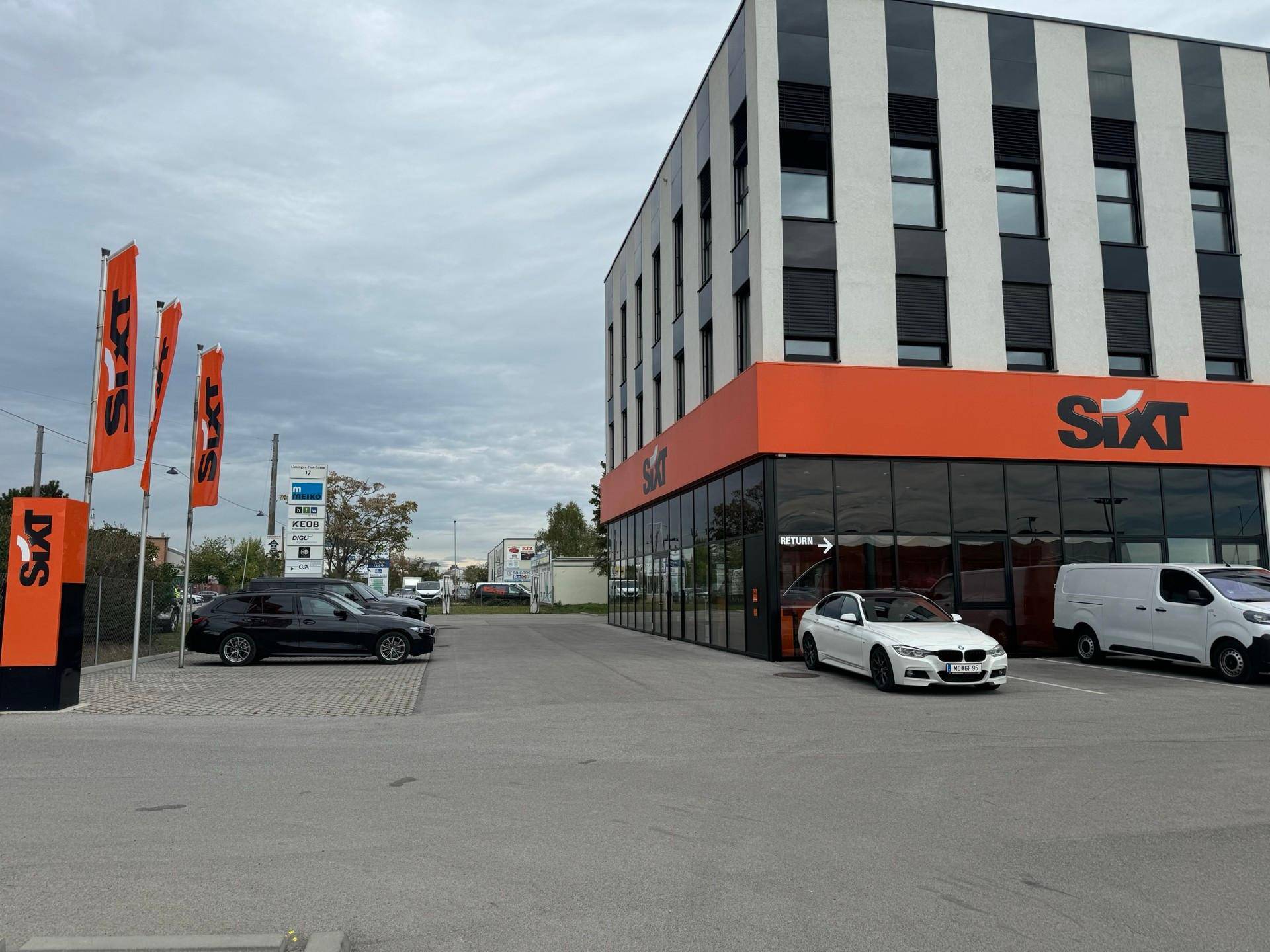 Außenansicht des modernen Geschäftsgebäudes mit Sixt-Filiale, Parkplätzen und Firmenflaggen unter bewölktem Himmel.