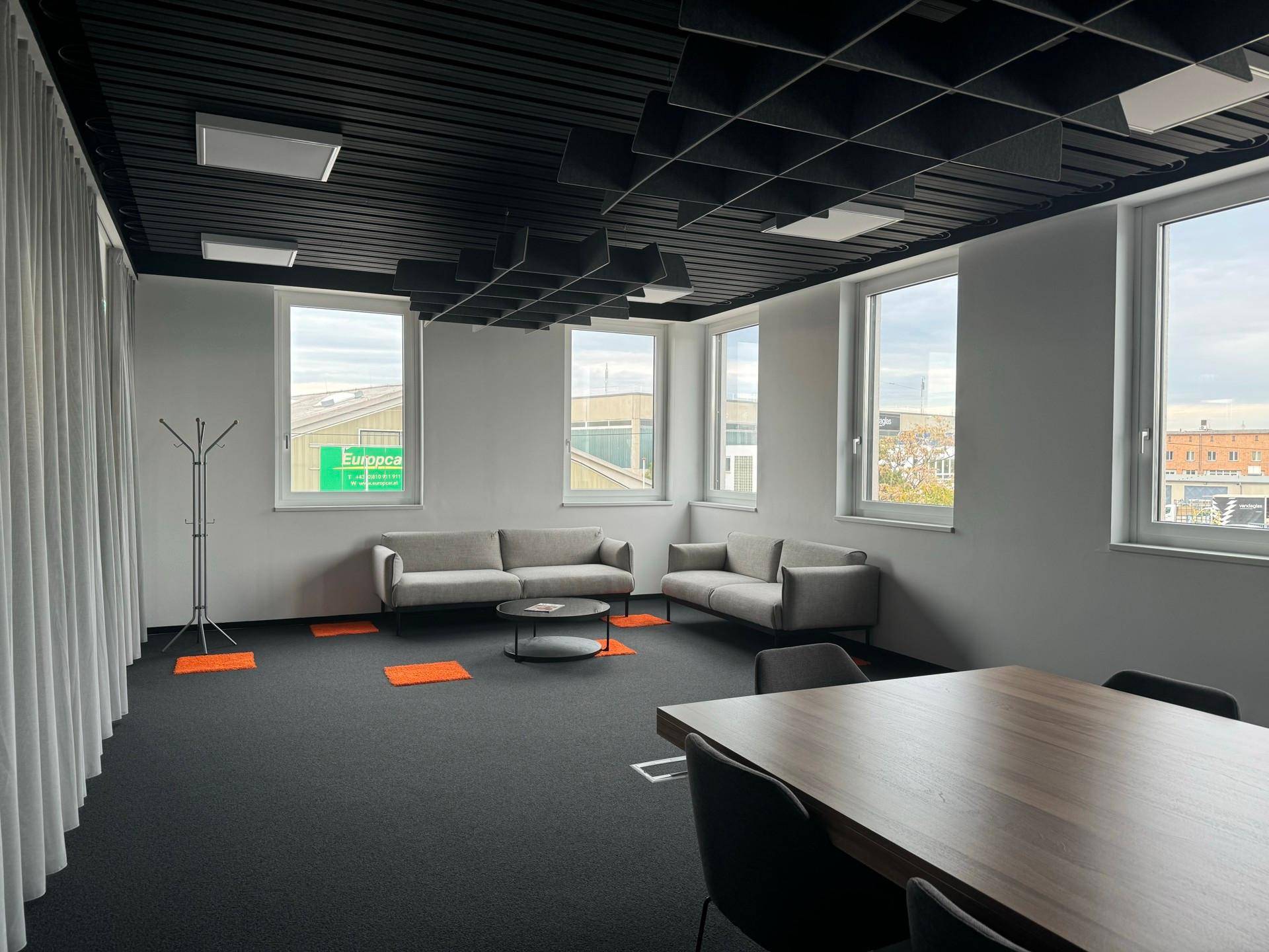 Moderner Büroloungebereich mit bequemen Sofas, Teppichboden und großen Fenstern, ergänzt durch akustische Deckenpaneele.