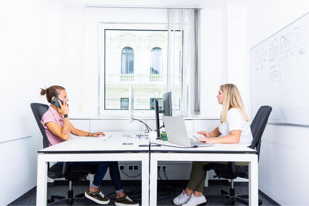 Zwei Personen arbeiten an hellen Schreibtischen in einem modernen Büro mit Blick auf die Stadt.