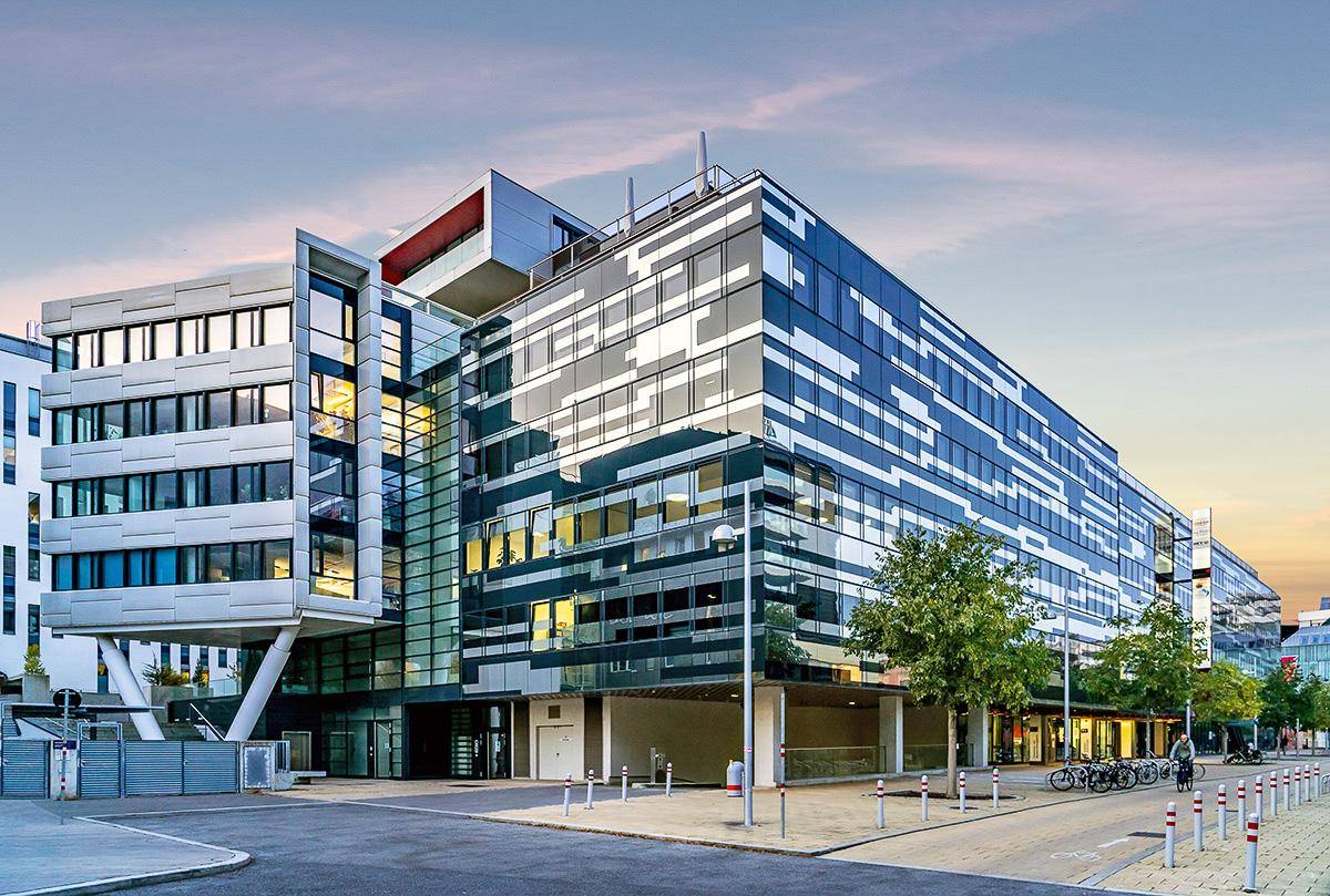 Architektonisch anspruchsvolles Gebäude mit Glasfassade und markanten horizontalen Elementen bei Sonnenuntergang, umgeben von städtischer Infrastruktur.