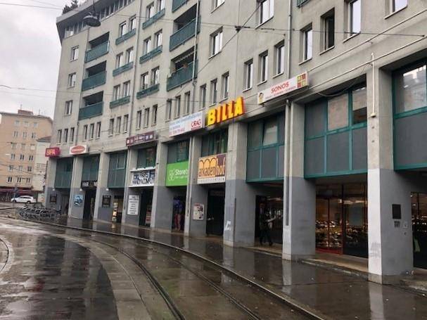 Mehrstöckiges Gebäude mit Geschäften im Erdgeschoss und Straßenbahnschienen im Vordergrund an einem regnerischen Tag.