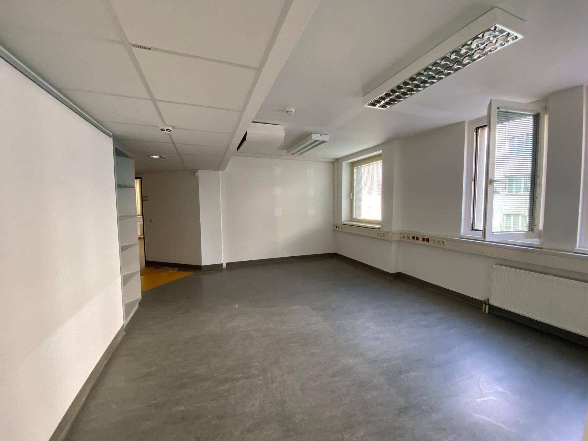 Geräumiger, leerer Büroraum mit abgehängter Decke, Fenstern und einem grauen Bodenbelag.
