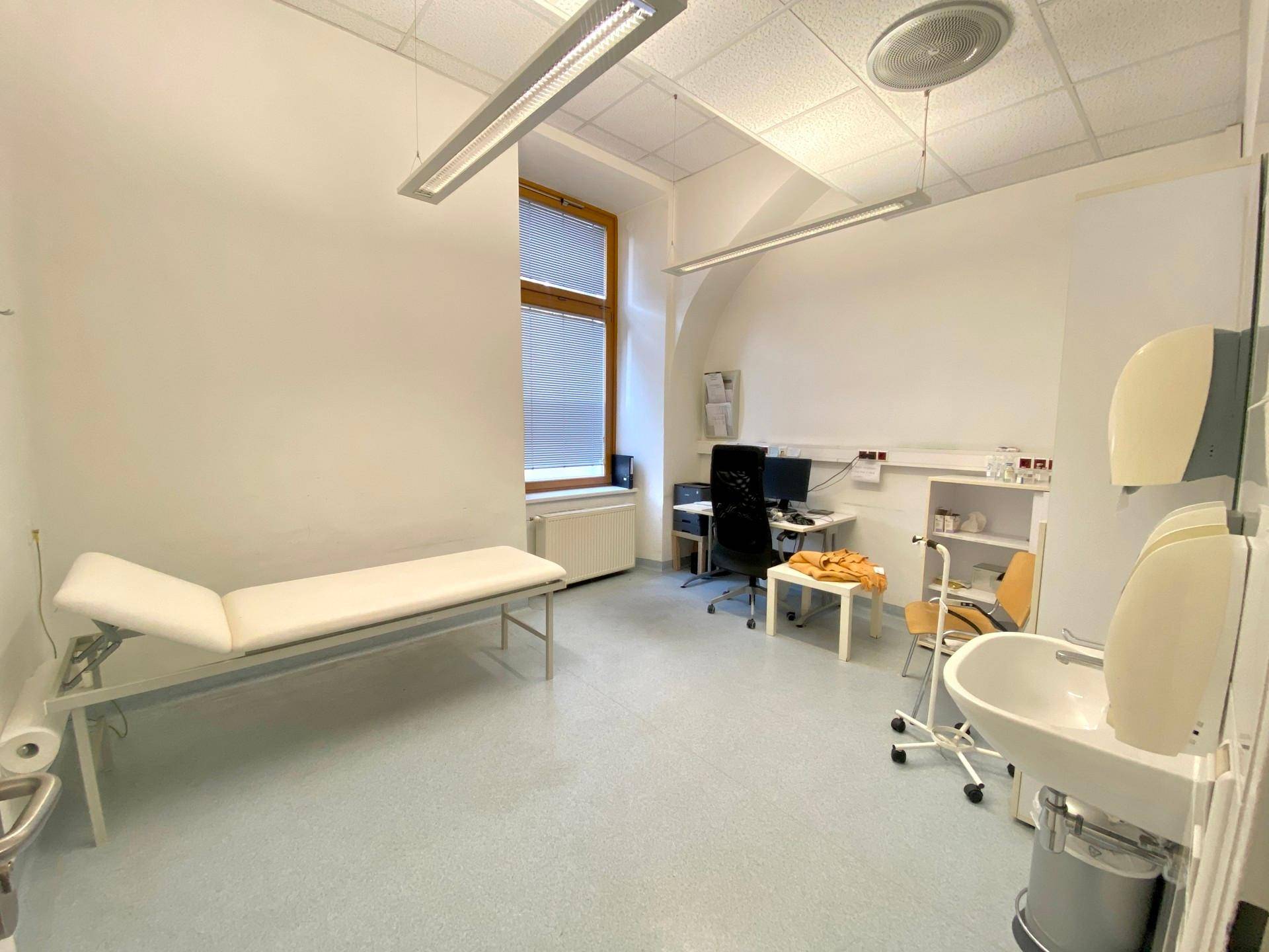 Heller Untersuchungsraum mit einem Behandlungsbett, medizinischer Ausstattung und großen Fenstern.