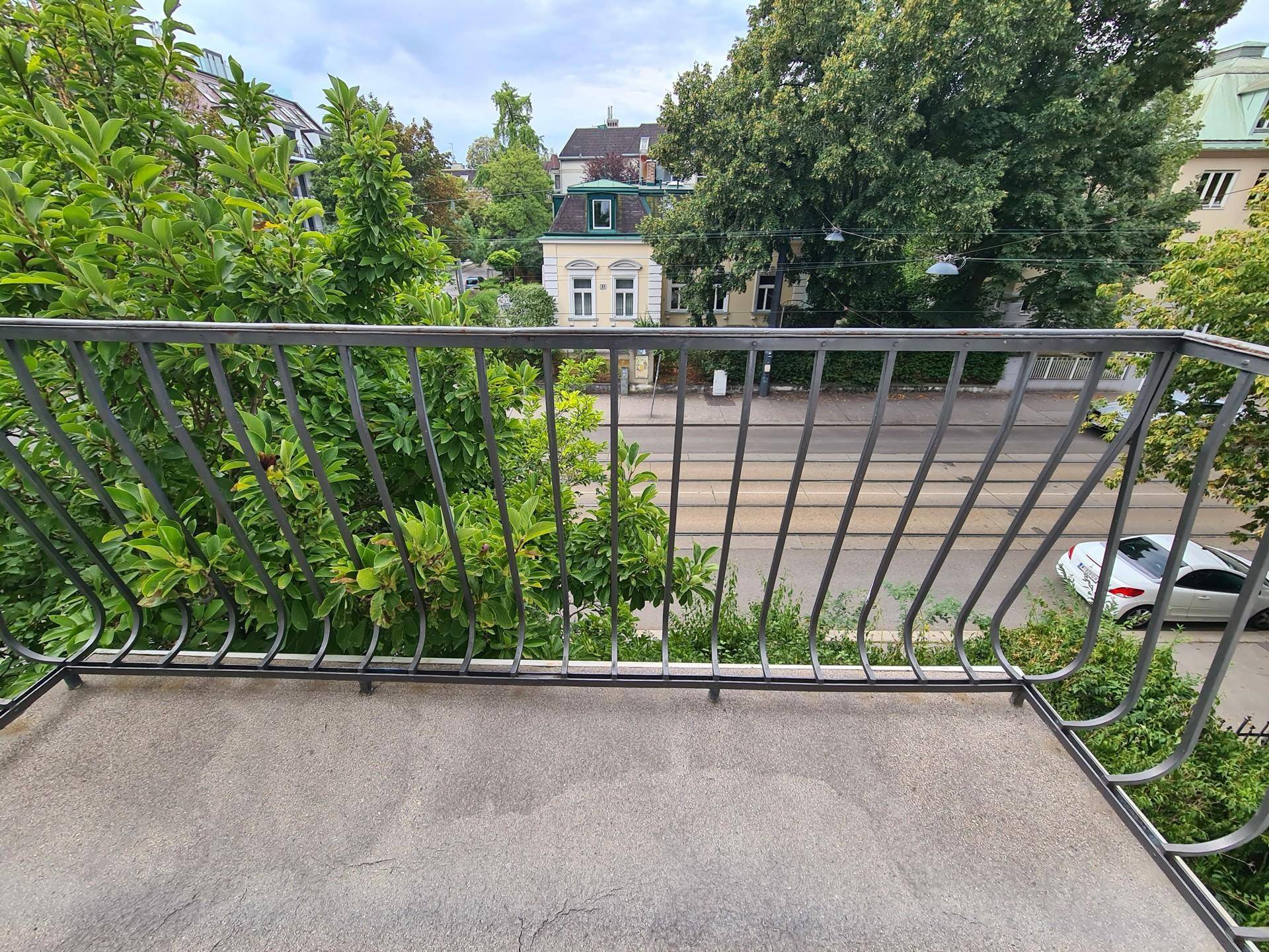 Blick vom Balkon auf die Straße und die gegenüberliegenden Gebäude, umgeben von Bäumen.