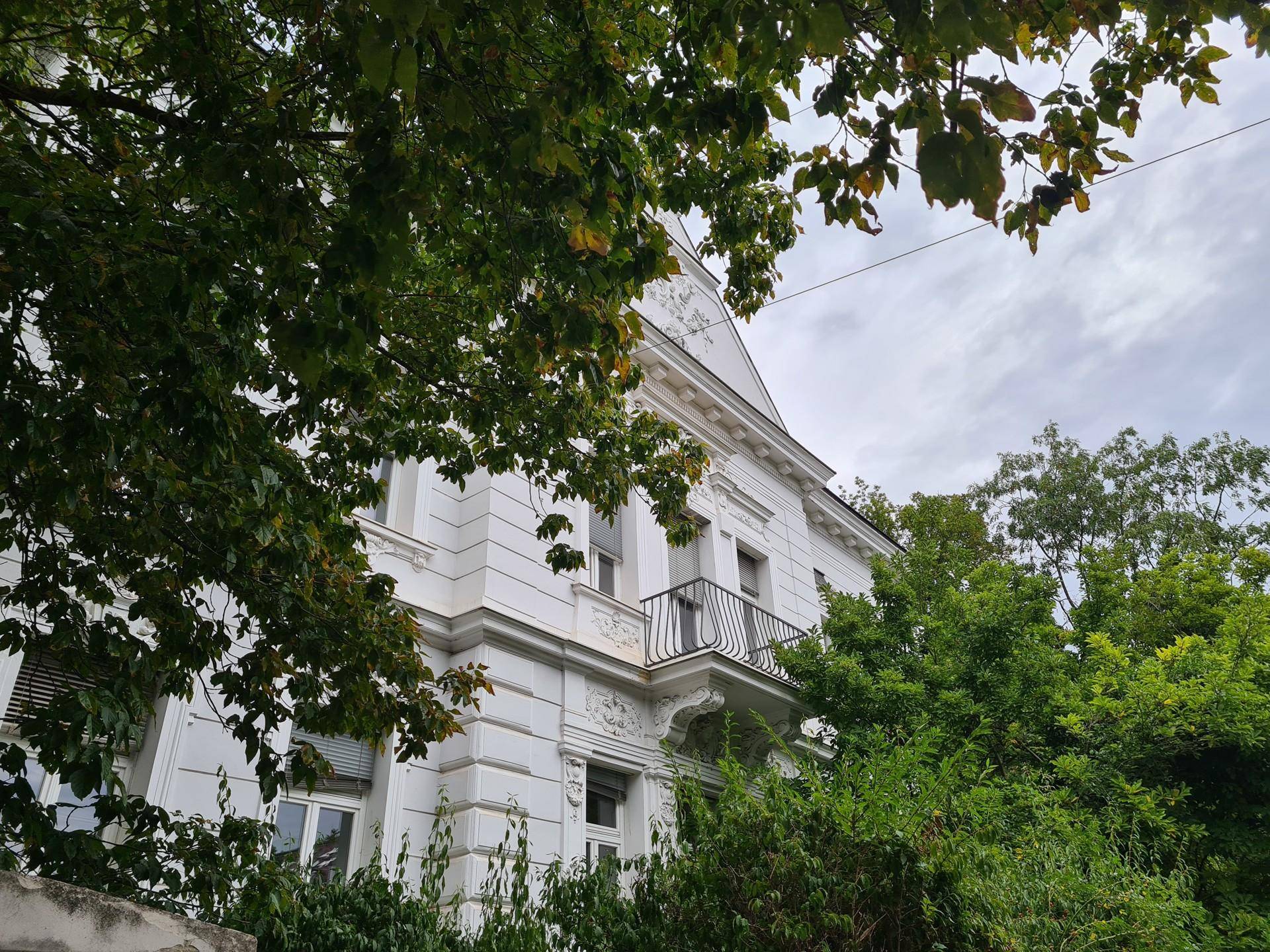 Seitenansicht des historischen Gebäudes mit Balkon, umgeben von dichtem Baumbestand.