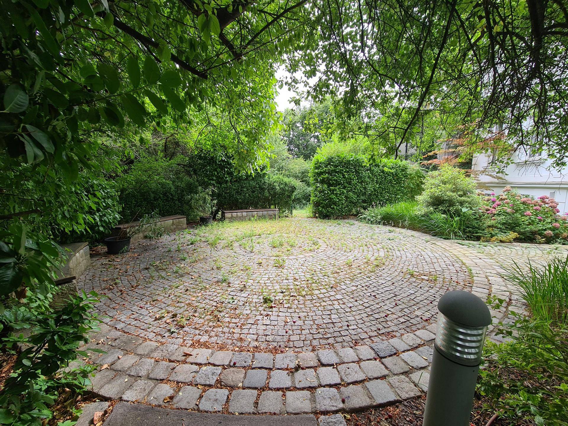 Gepflasterter Gartenbereich mit kreisförmigem Muster, umgeben von dichtem Grün und Bäumen.