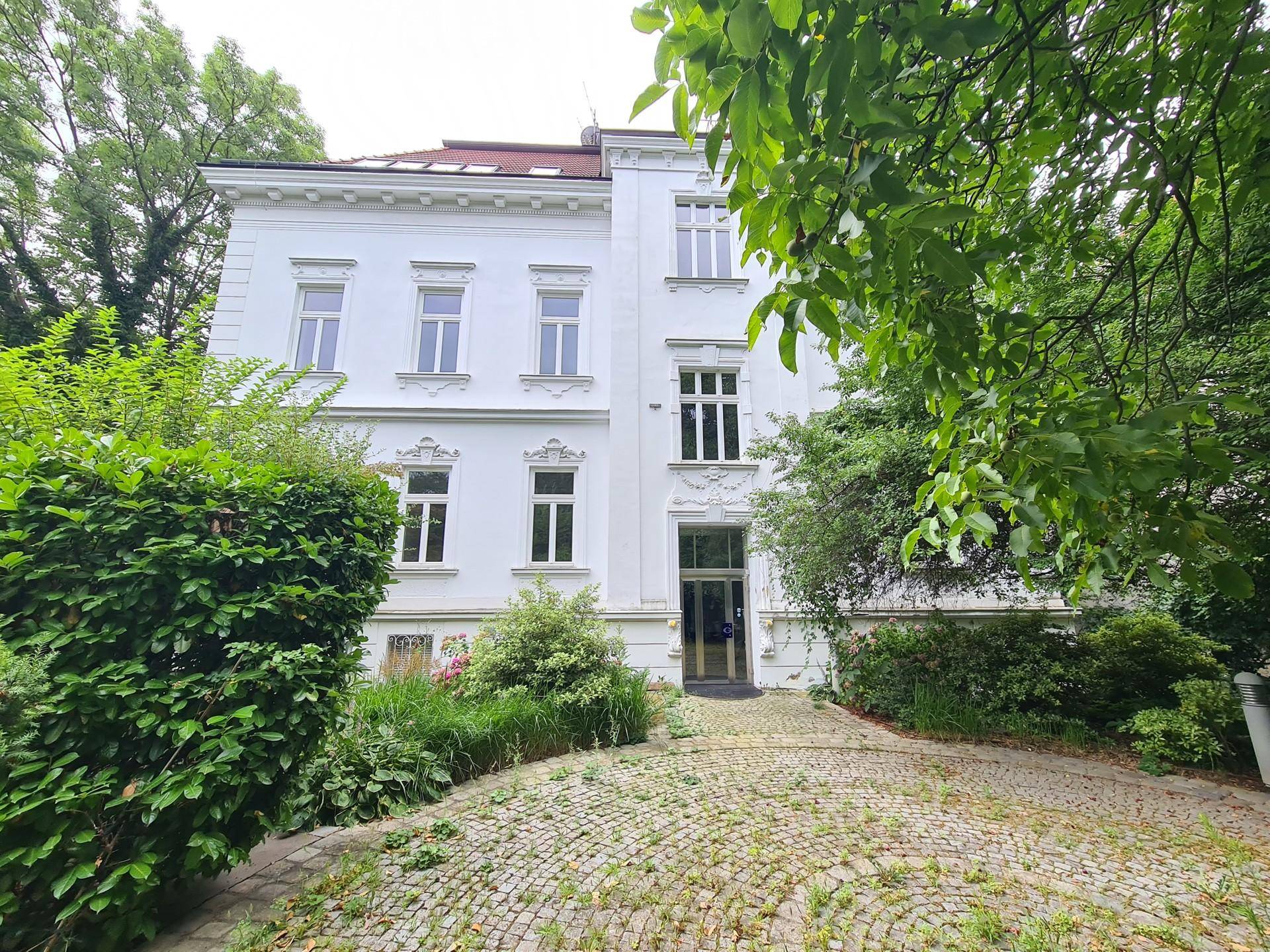 Historisches Gebäude mit gepflasterter Auffahrt, umgeben von üppigem Grün und Bäumen.