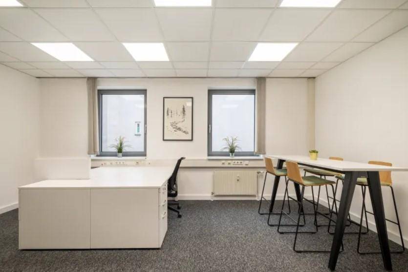 Funktionales Büro mit Teppichboden, einem Schreibtisch und einem Stehtischbereich unter zwei Fenstern.