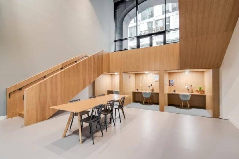 Moderner Bürobereich mit Holzakzenten, Gemeinschaftstisch, individuellen Arbeitskabinen und hohen Fenstern.
