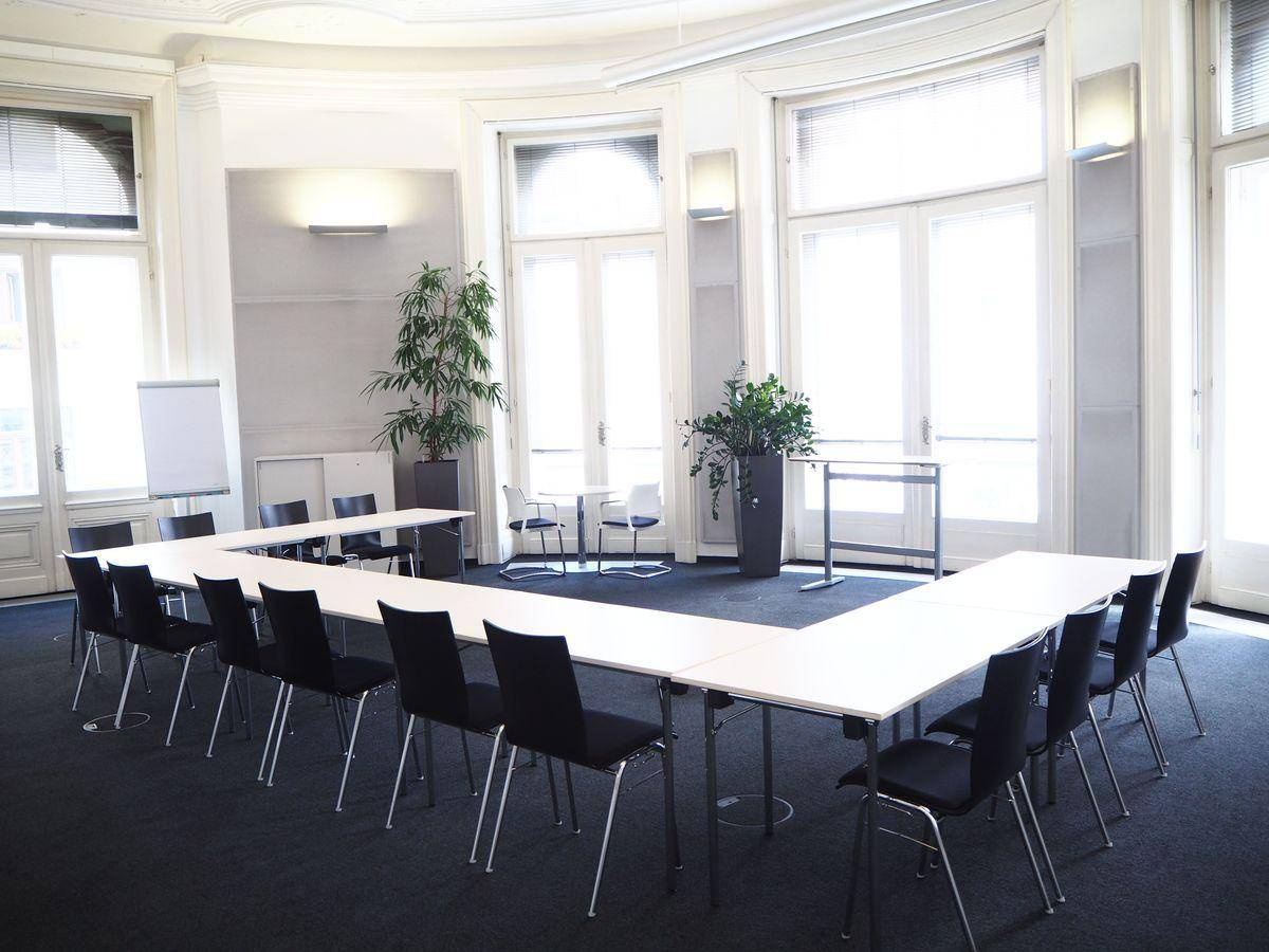 Großer Konferenzraum mit U-förmiger Tischordnung, vielen Stühlen und hohen Fenstern.