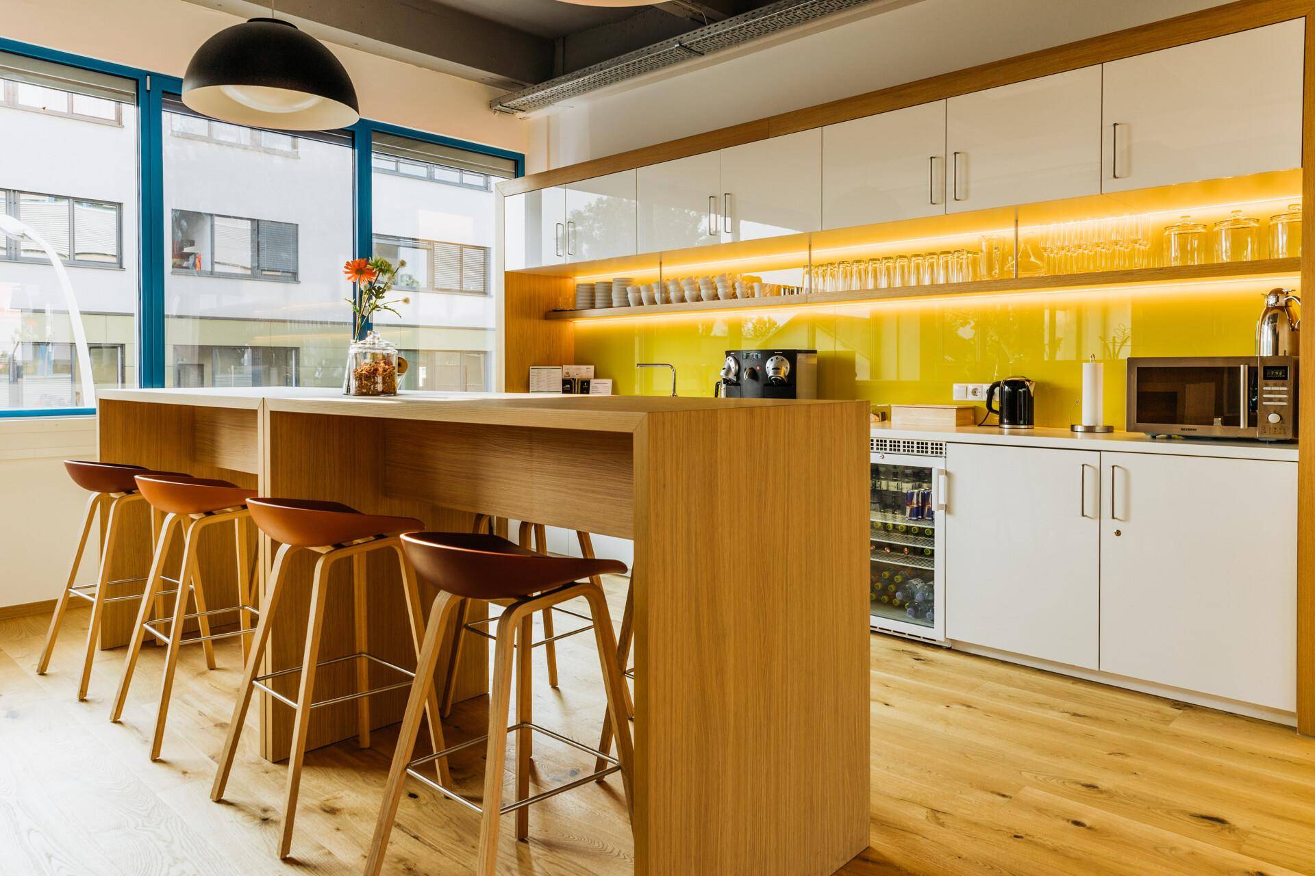 Moderne Teeküche mit Holztresen, Barhockern, weißen Schränken und gelber Akzentwand, perfekt für Pausen.
