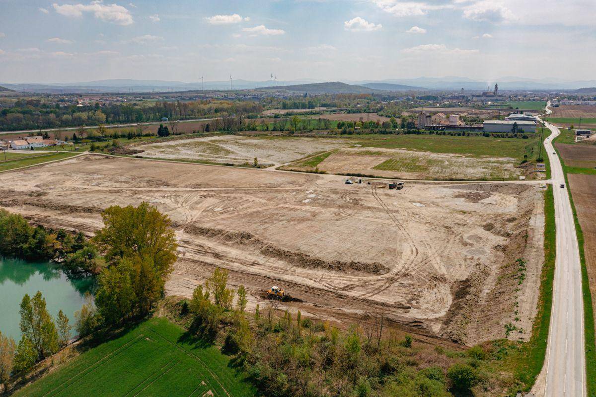 Weitläufige Baustelle mit Erdarbeiten und einem Bagger, angrenzend an einen See und eine Landstraße.