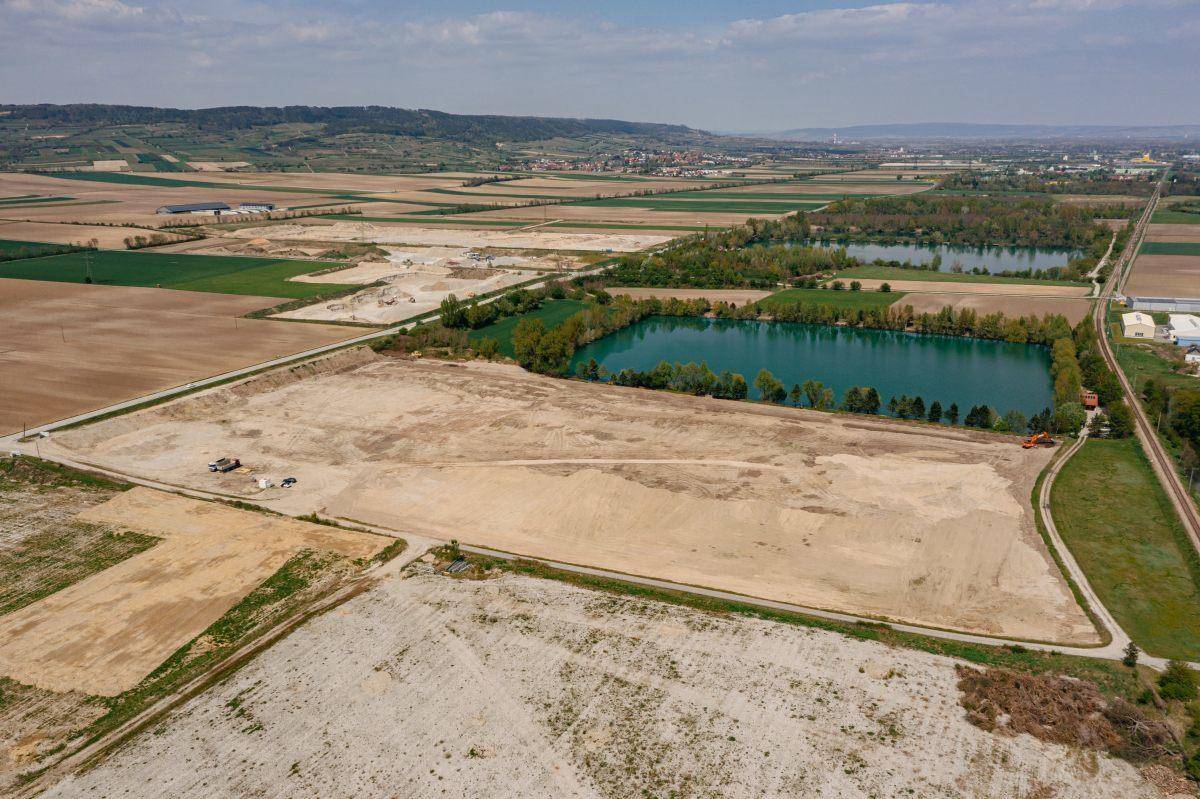 Luftbild einer weitläufigen Baustelle neben einem See, umgeben von landwirtschaftlichen Flächen und Natur.