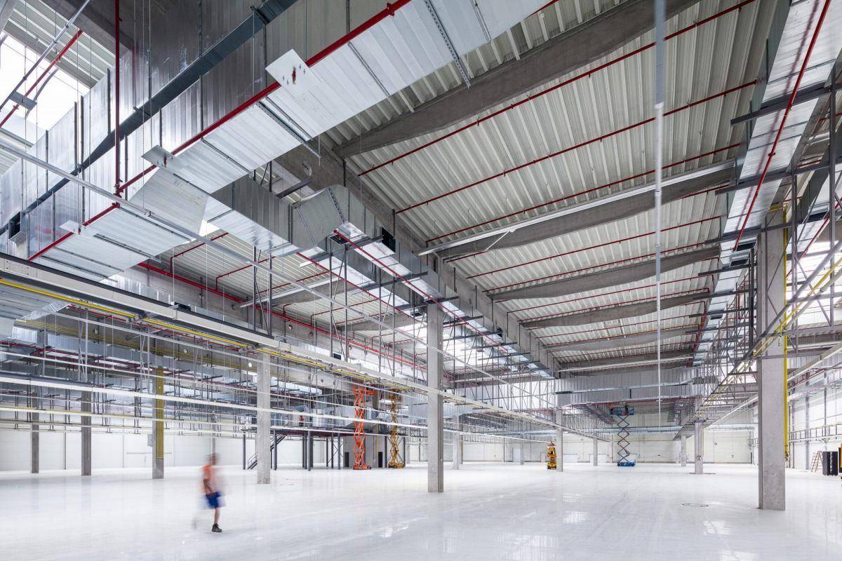 Geräumige, leere Lagerhalle im Bau mit hohen Decken und sichtbaren Lüftungsanlagen und Installationen.