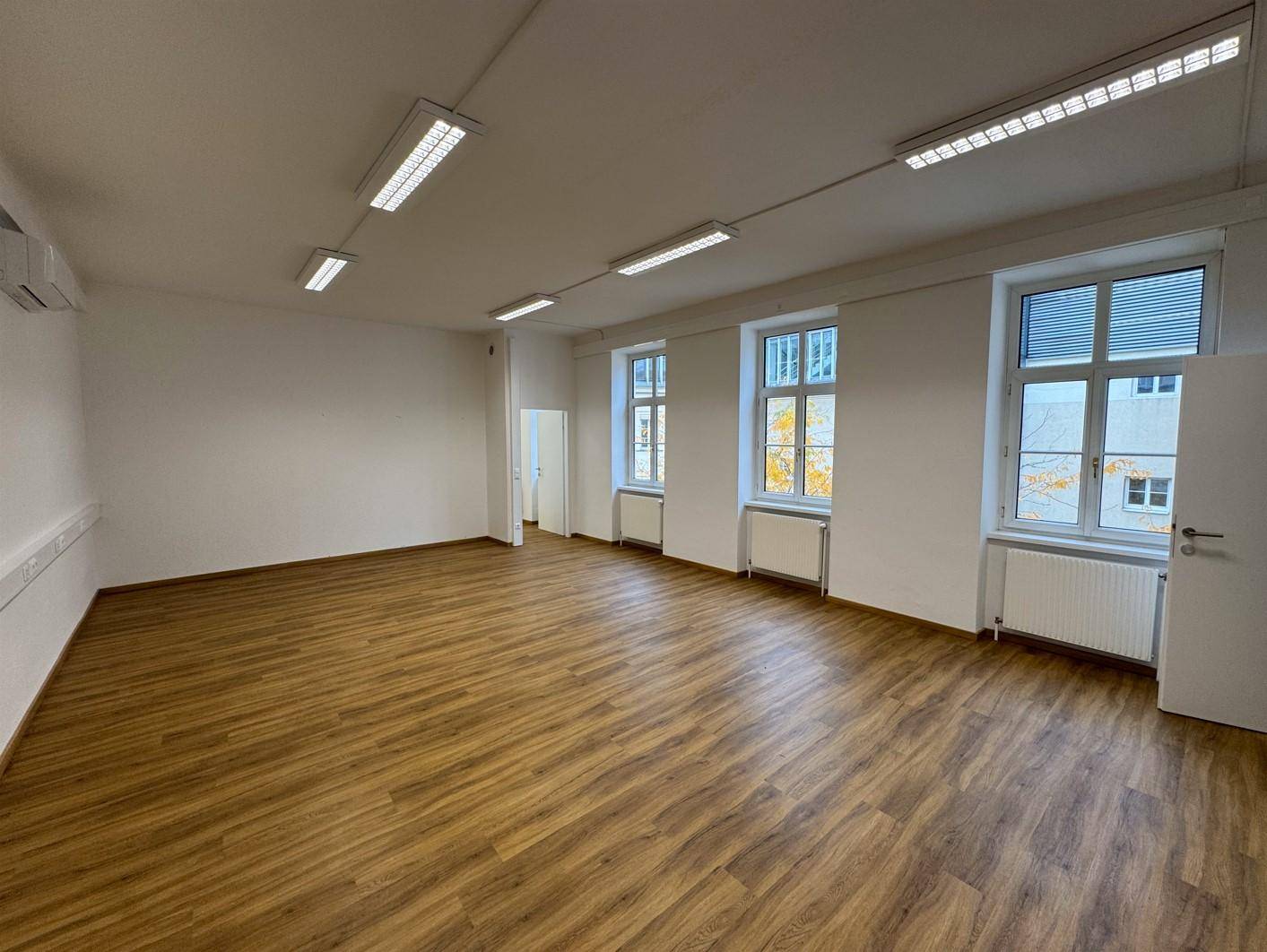 Geräumiges, leeres Büro mit großen Fenstern, Holzfußboden und moderner Deckenbeleuchtung.