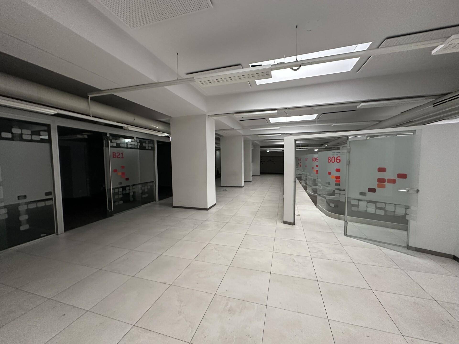 Langer Flur in einem Bürogebäude mit weißen Fliesen, Glaswänden und sichtbaren Lüftungsrohren an der Decke.