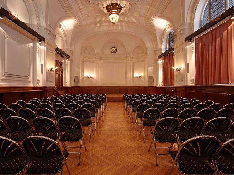 Großer Veranstaltungssaal mit Reihenbestuhlung, elegantem Holzboden und hohen, verzierten Decken.