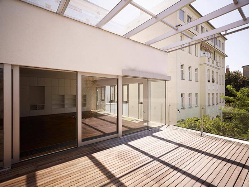 Große Dachterrasse mit Holzbelag und einer modernen Glasüberdachung, angrenzend an helle Innenräume.