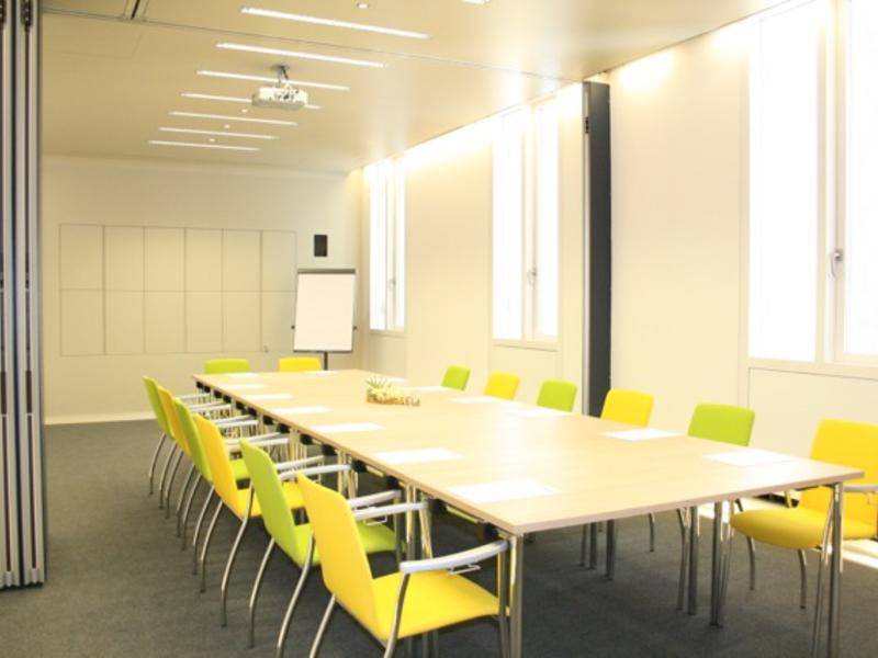 Heller Konferenzraum mit langem Tisch, vielen bunten Stühlen und großen Fenstern für Tageslicht.