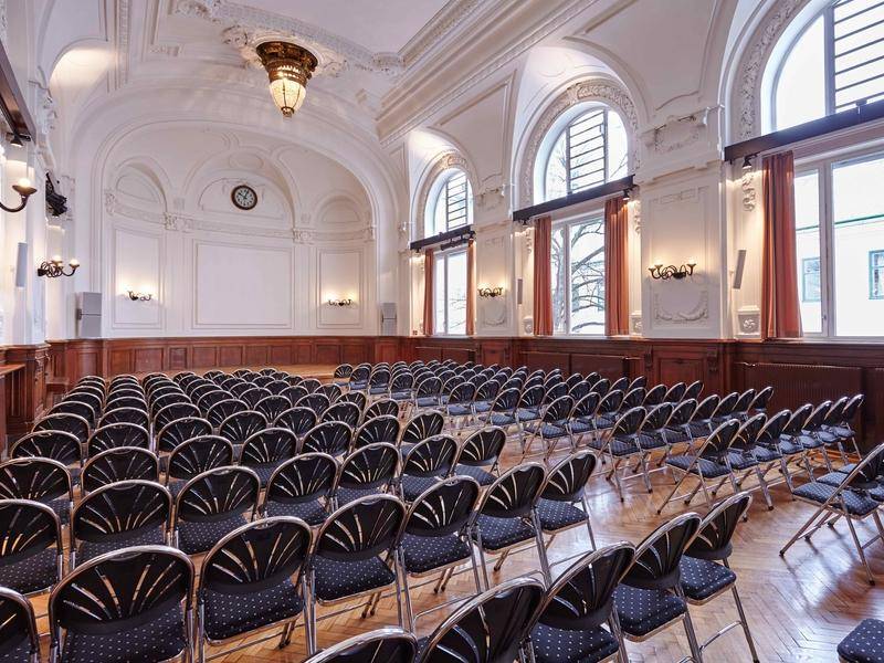 Geräumiger Veranstaltungssaal mit eleganten Stühlen, Parkettboden und großen Bogenfenstern, die viel Licht spenden.