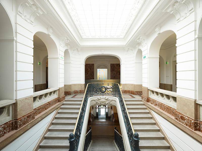 Prunkvolles Treppenhaus mit Marmorverkleidung, kunstvollem Geländer und einem hellen Oberlicht an der Decke.