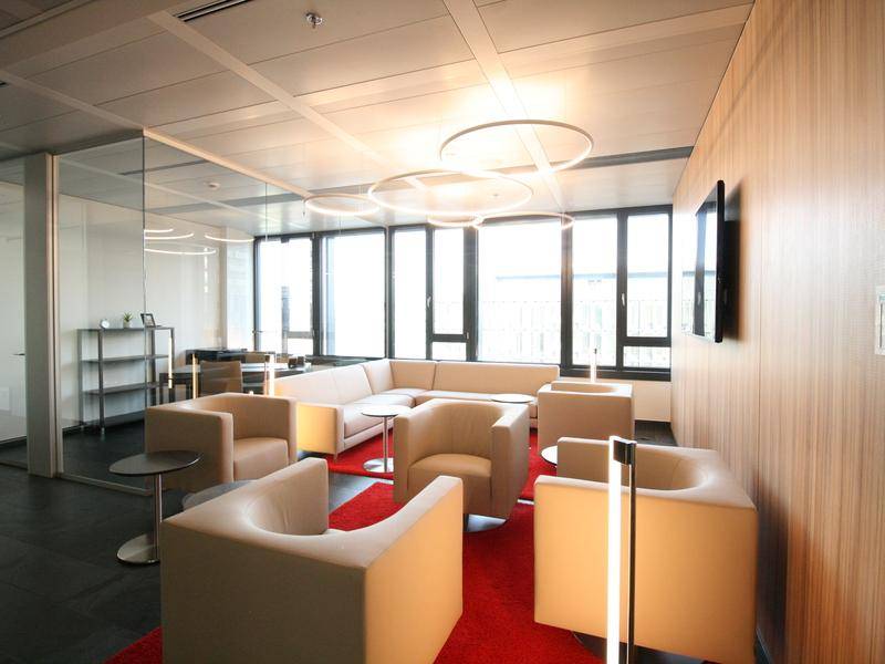 Elegante Lounge mit beigen Sitzmöbeln, roten Teppichen und großen Fenstern, die eine helle und einladende Atmosphäre schaffen.