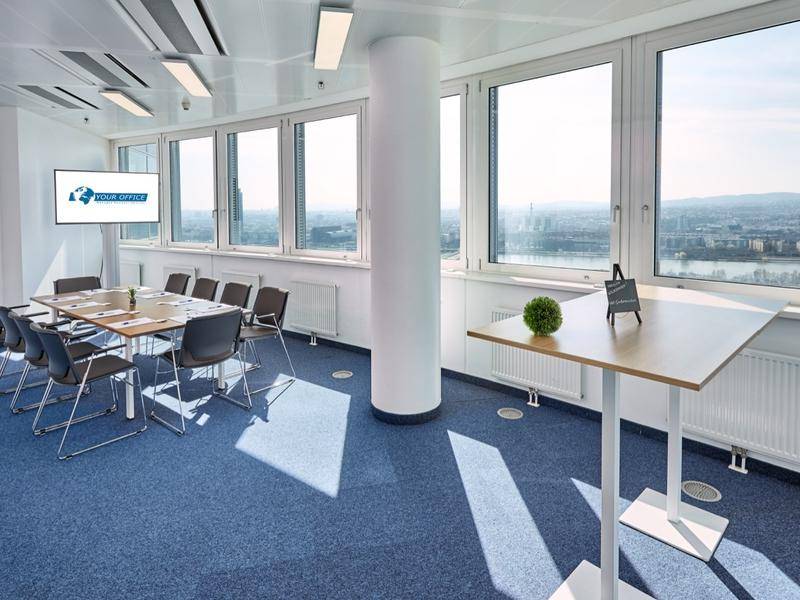 Heller Konferenzraum mit blauem Teppich, großem Fenster und Blick auf die Stadt.