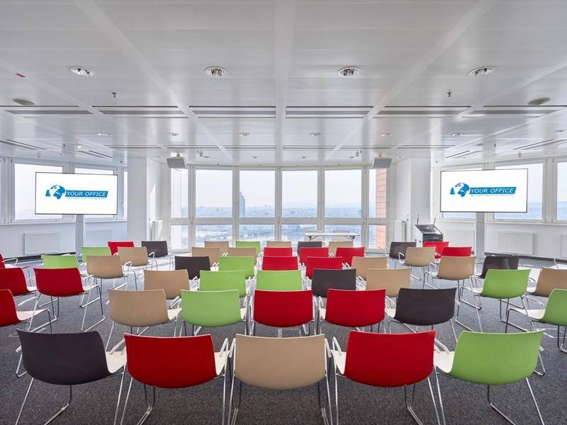 Großer Konferenzraum mit Reihenbestuhlung, zwei Bildschirmen und weitem Stadtblick.