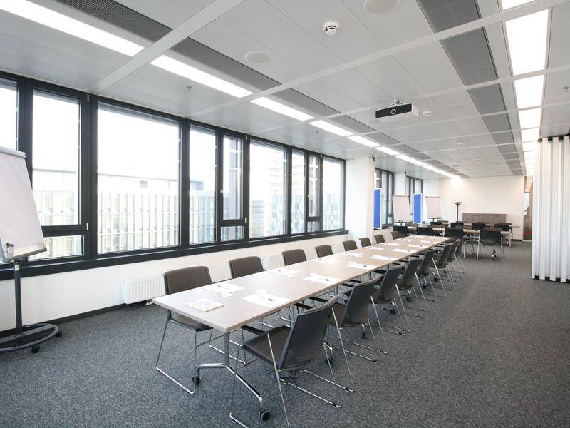Heller Konferenzraum mit mehreren Tischen, bequemen Stühlen und einer langen Fensterfront, die den Raum erhellt.