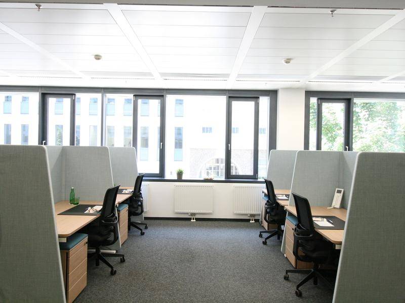 Offener Bürobereich mit mehreren Arbeitsplätzen, Trennwänden und großen Fenstern.
