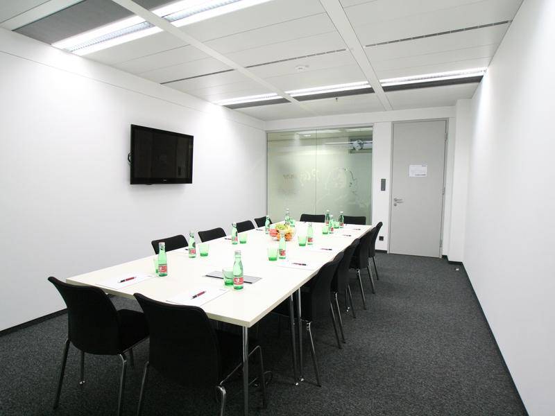 Heller Konferenzraum mit großem Tisch, schwarzen Stühlen und einem Wandfernseher.