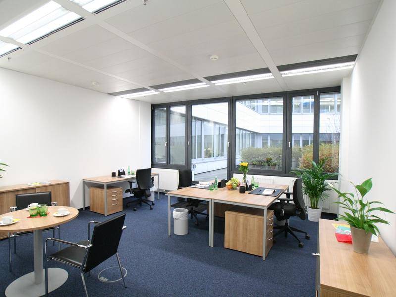 Helles Büro mit mehreren Arbeitsplätzen, blauer Teppichboden und Pflanzen.