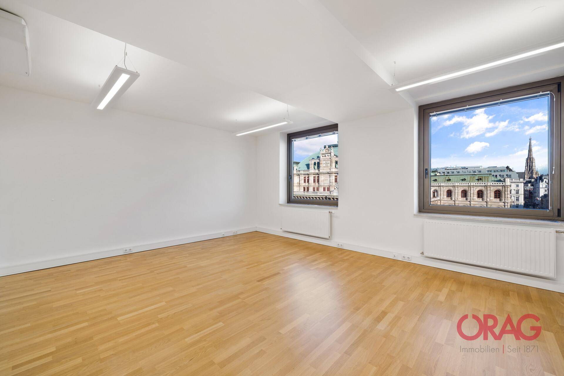 Leeres Büro mit hellem Holzboden, weißen Wänden und Fenstern, die einen schönen Stadtblick bieten.
