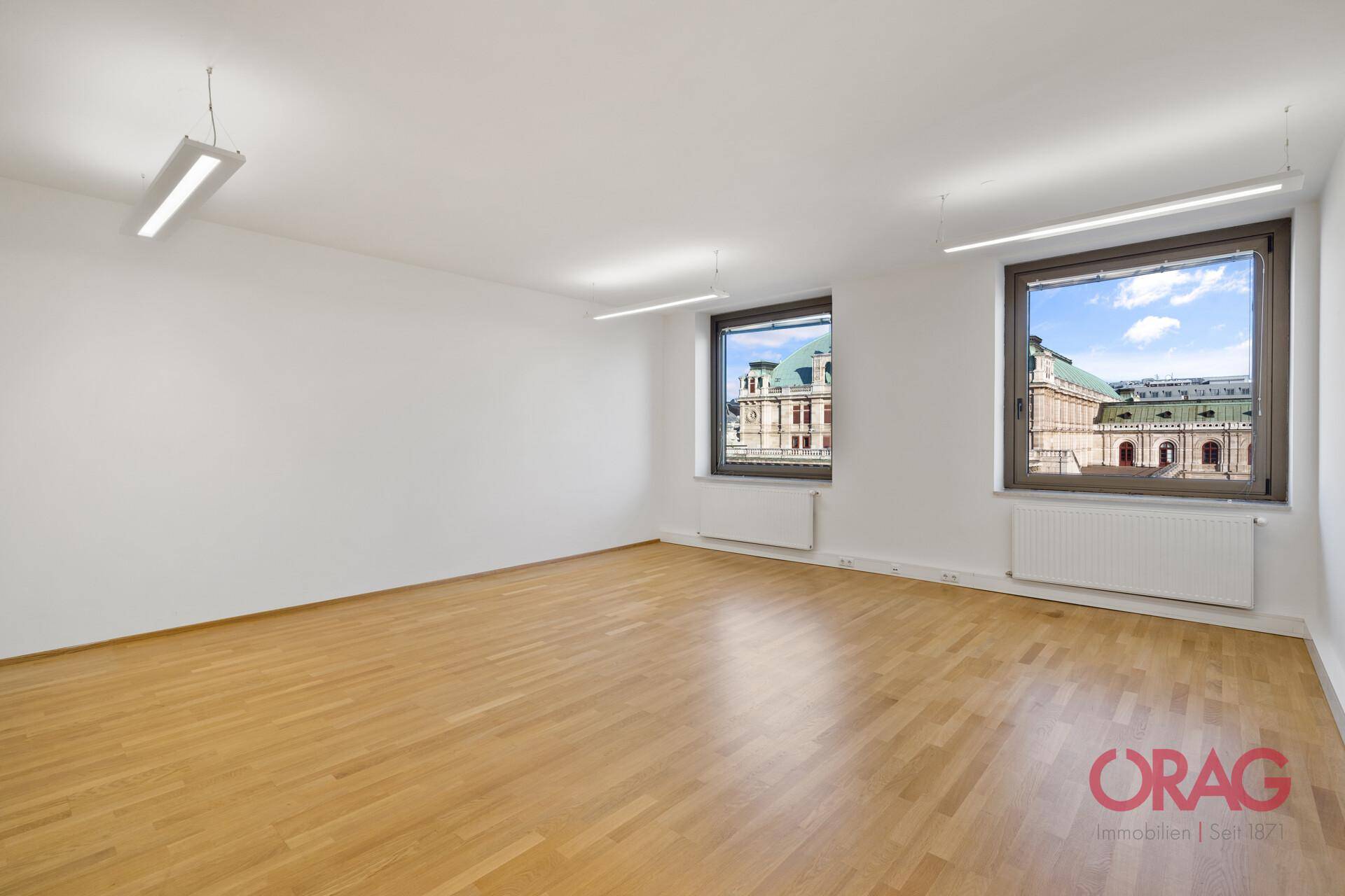 Leeres Büro mit hellem Holzboden, weißen Wänden und Fenstern mit Blick auf die Stadt.