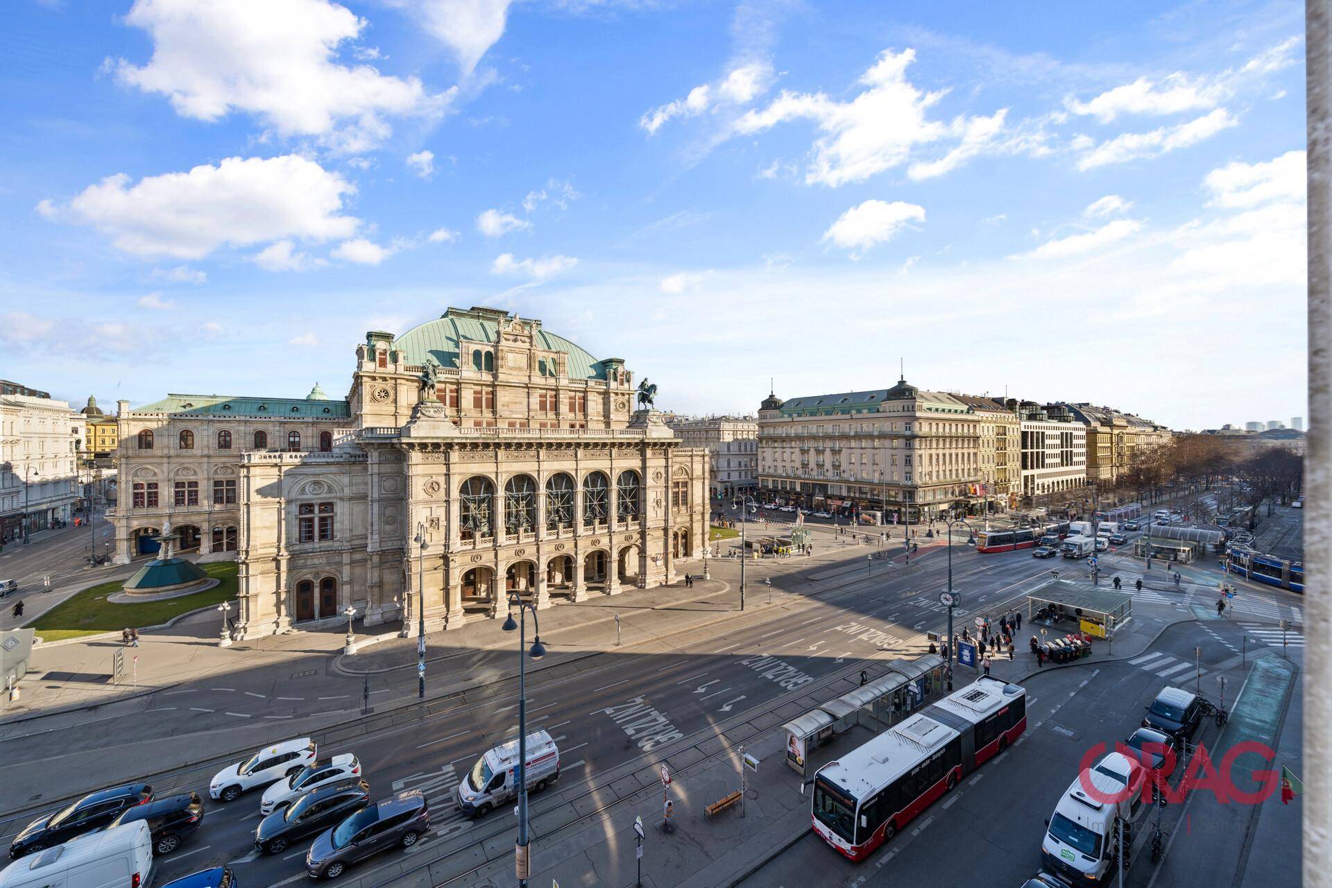 Panoramablick auf die Wiener Staatsoper und die belebte Kreuzung an einem sonnigen Tag mit blauem Himmel.