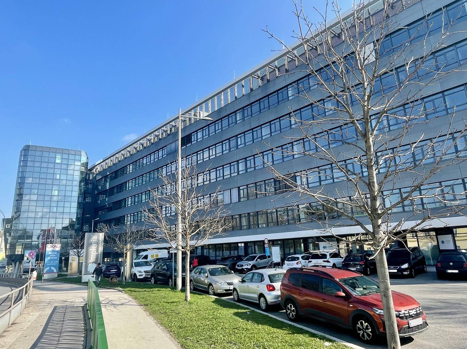 Moderne Bürogebäude mit vielen Fenstern und geparkten Autos auf dem Vorplatz unter blauem Himmel.