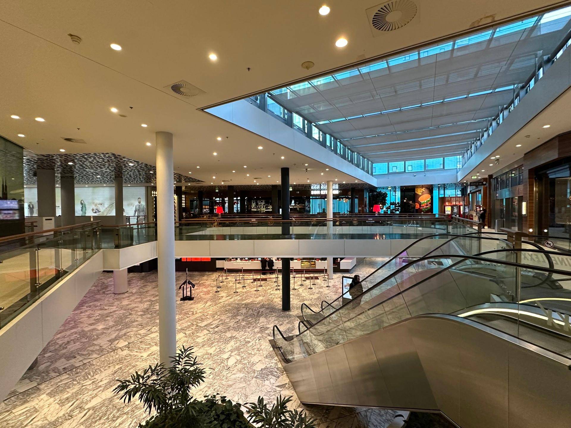 Helles, modernes Einkaufszentrum mit Rolltreppen, Marmorboden und viel Tageslicht durch das Glasdach.
