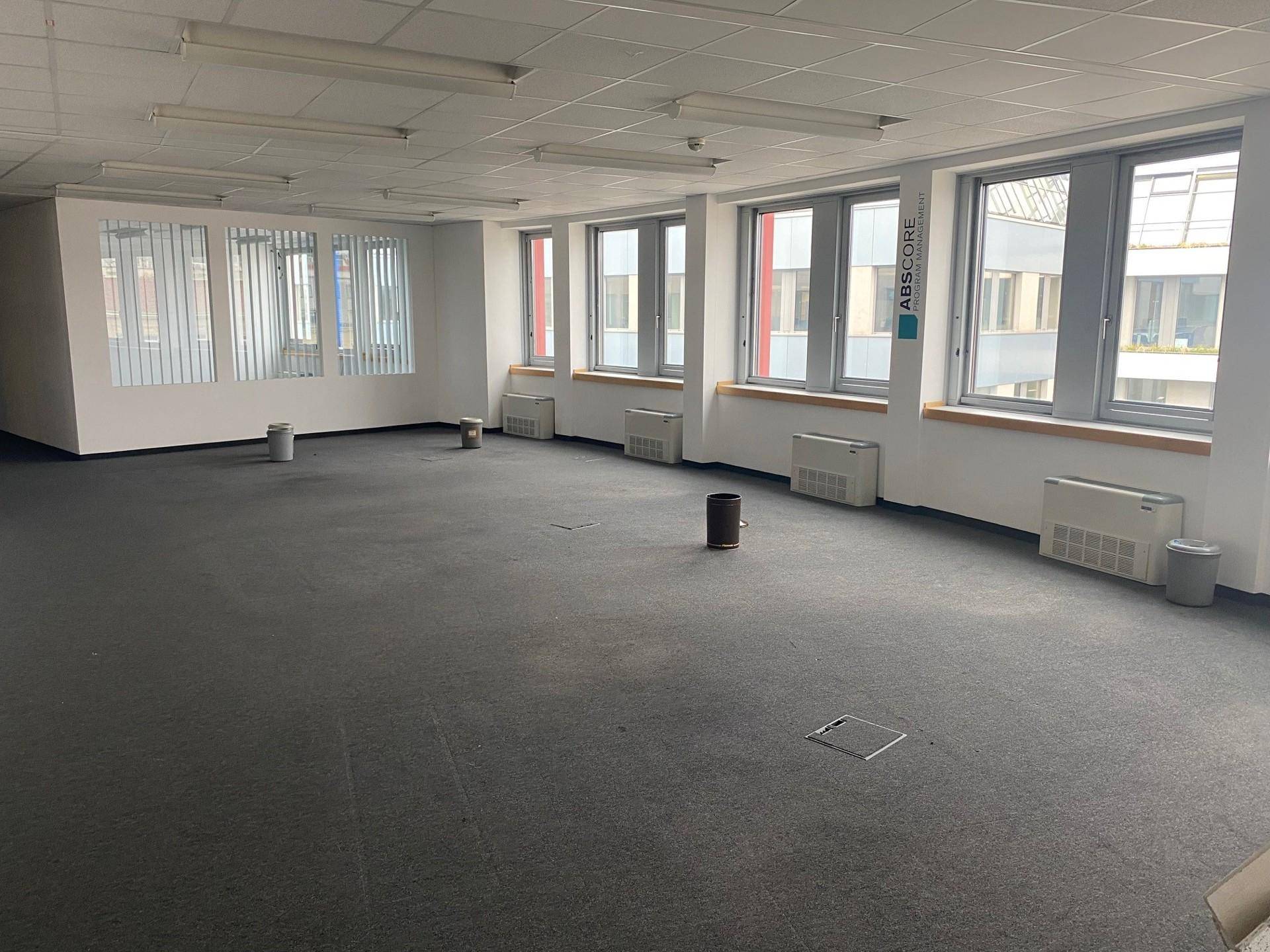 Weitläufiges, leeres Büro mit grauem Teppichboden, vielen Fenstern und Deckenbeleuchtung, mit Blick auf die Stadt.