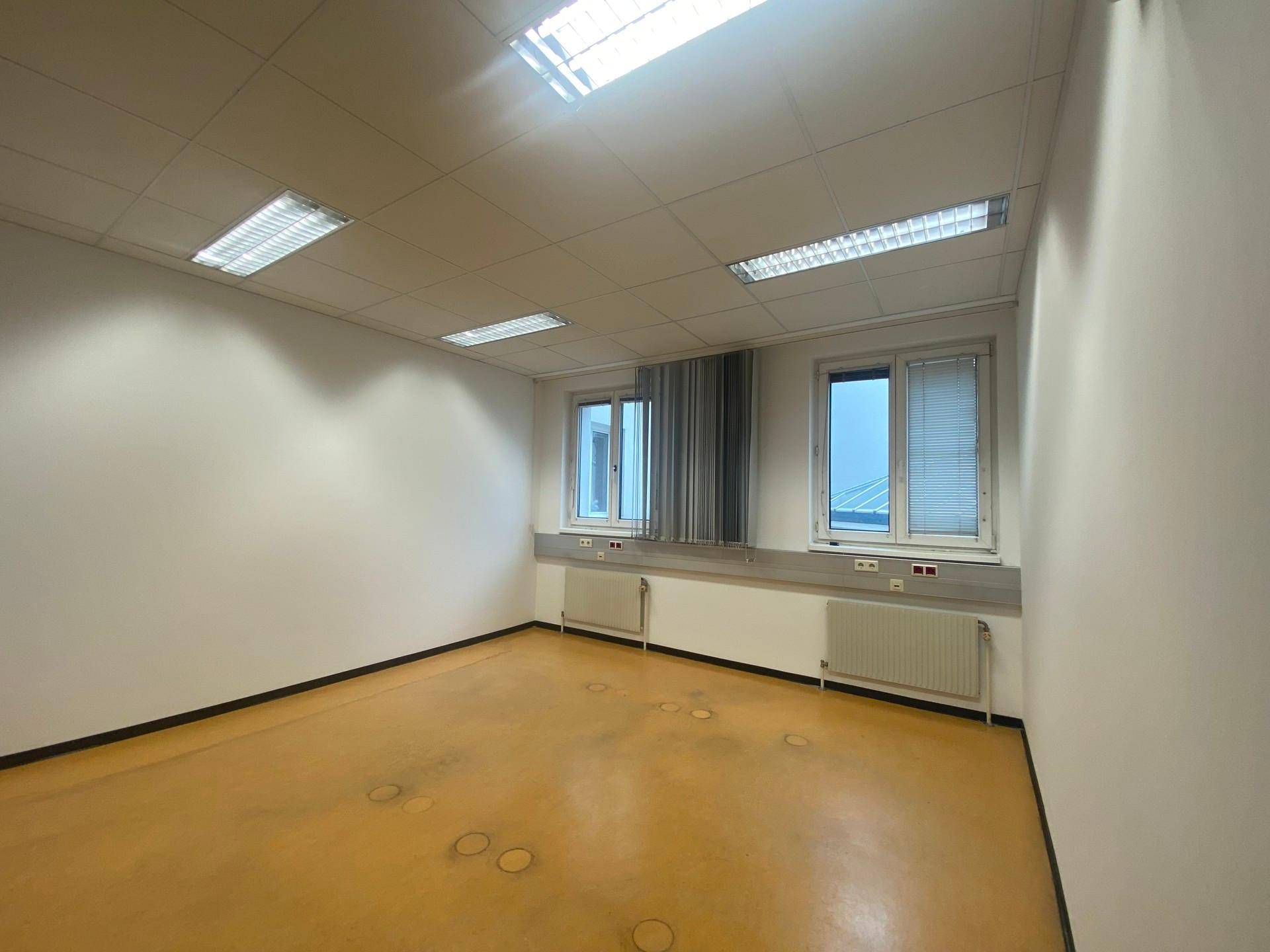 Kleinerer, leerer Büroraum mit gelbem Bodenbelag, zwei Fenstern und effizienter Deckenbeleuchtung.