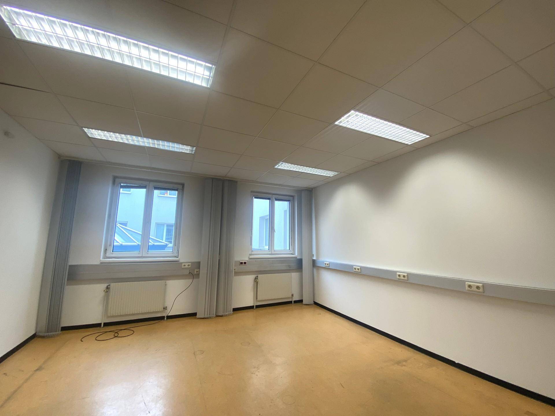 Leerer Büroraum mit gelbem Bodenbelag, zwei Fenstern und praktischen Wandsteckdosenleisten.