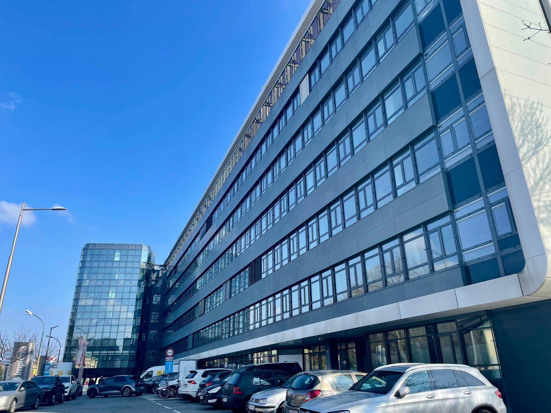 Modernes Bürogebäude mit Glasfassade und zahlreichen Fenstern, umgeben von einem Parkplatz unter blauem Himmel.