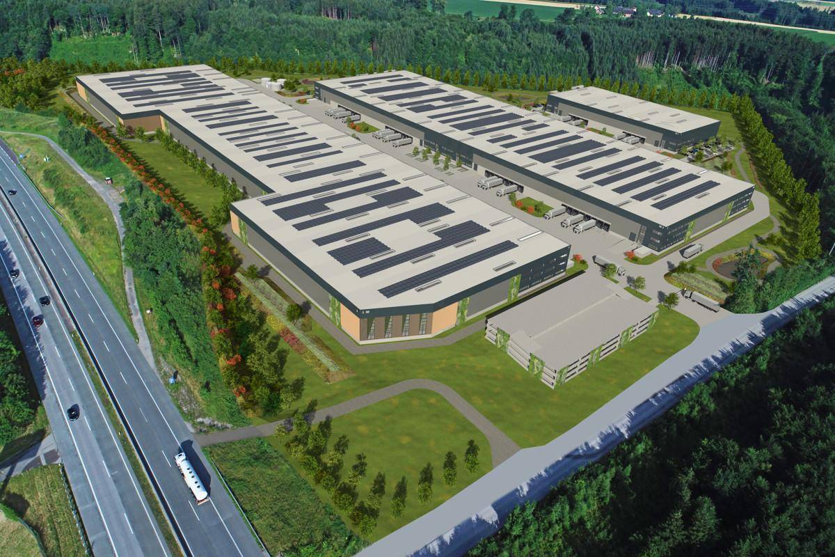 Weitläufige Luftaufnahme eines Industrieparks mit Solaranlagen auf den Dächern, angrenzend an eine Autobahn und grüne Wälder.