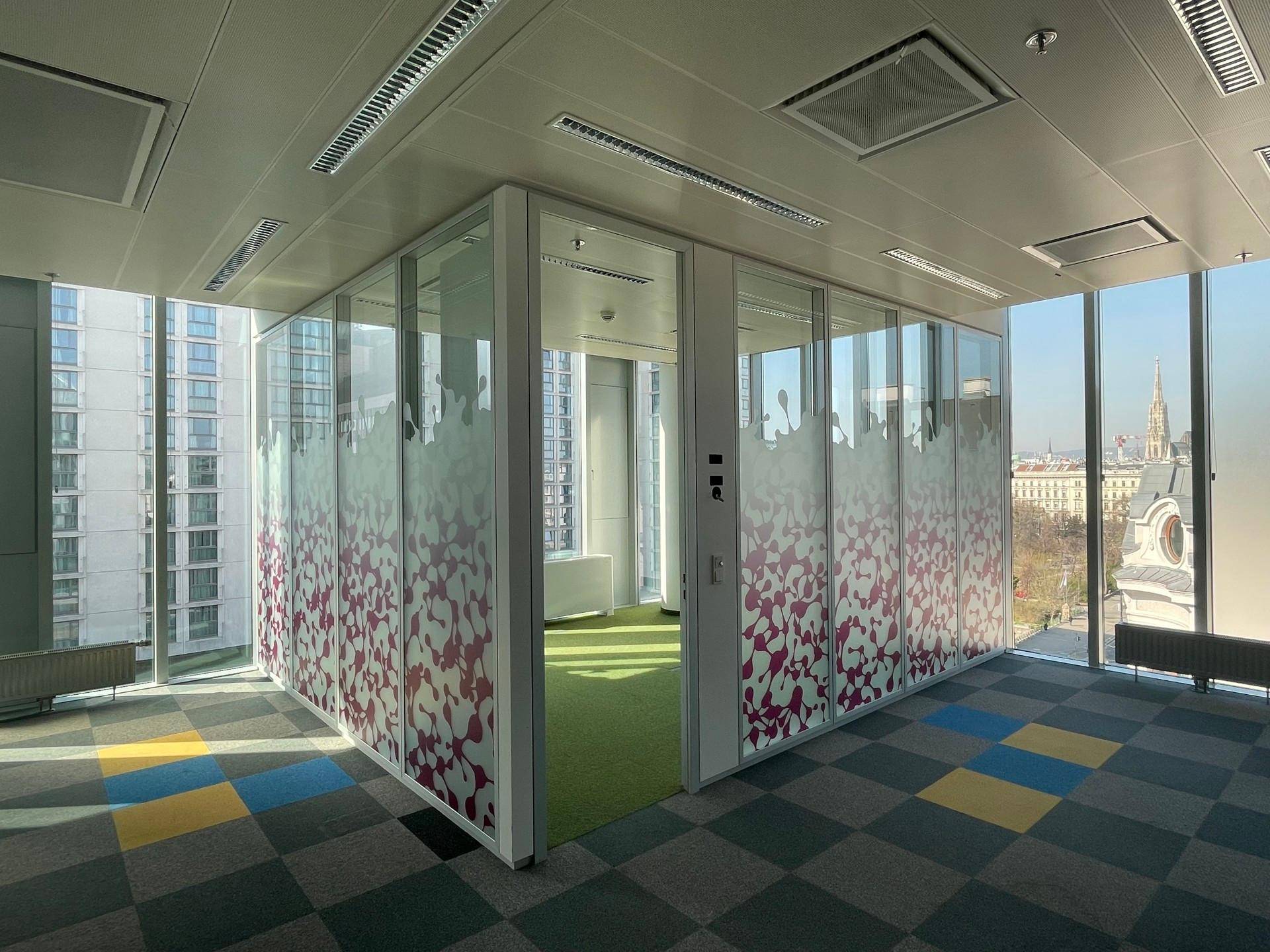Moderne Büroräume mit Glaswänden, grünem Teppich und Blick auf die Stadt.