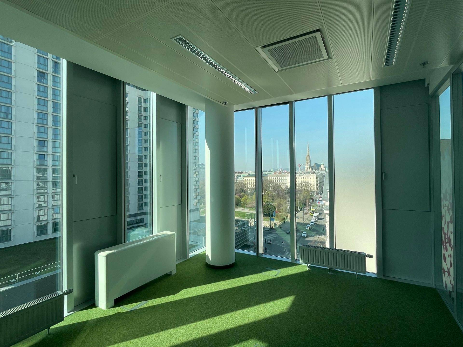 Helles Büro mit grünem Teppichboden und raumhohen Fenstern, die einen weiten Blick über die Stadt bieten.