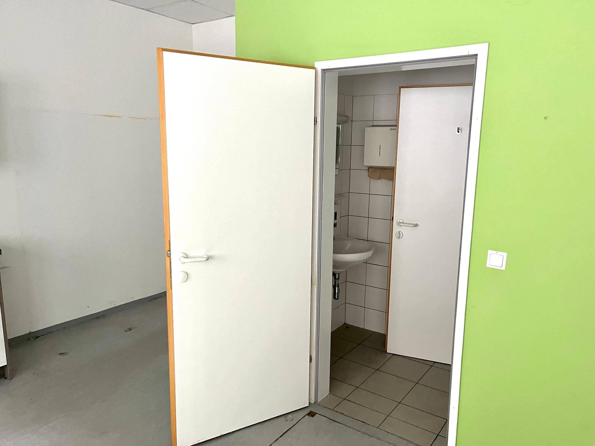 Blick in einen funktionalen Toilettenraum mit Waschbecken und weißen Fliesen, angrenzend an einen leeren Raum mit grüner Wand.