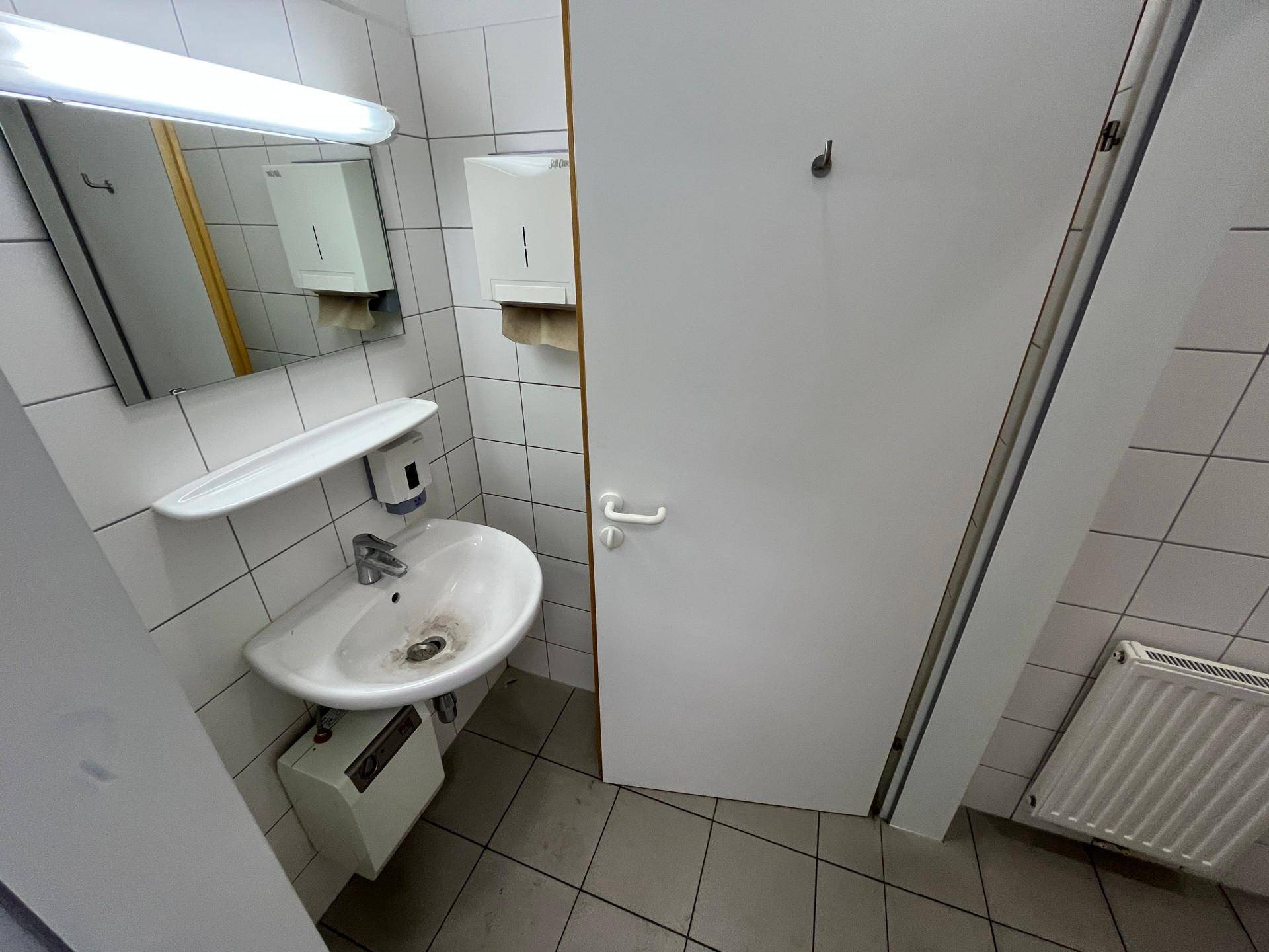 Kleiner Toilettenraum mit weißen Fliesen, Waschbecken und Spiegel, funktional und sauber gehalten.