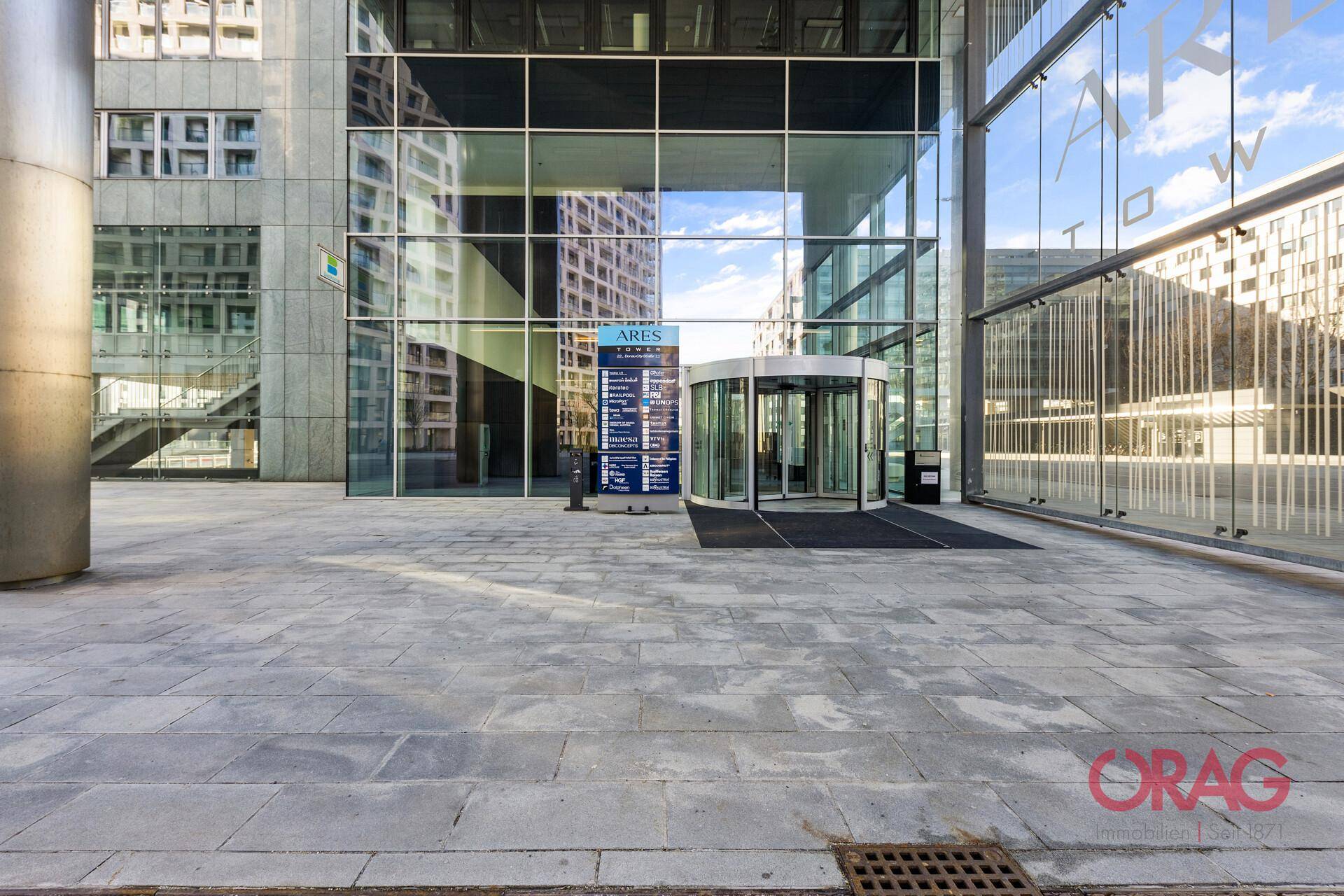 Moderner Gebäudeeingang mit Drehtür und weitläufiger Glasfassade, umgeben von Pflastersteinen.