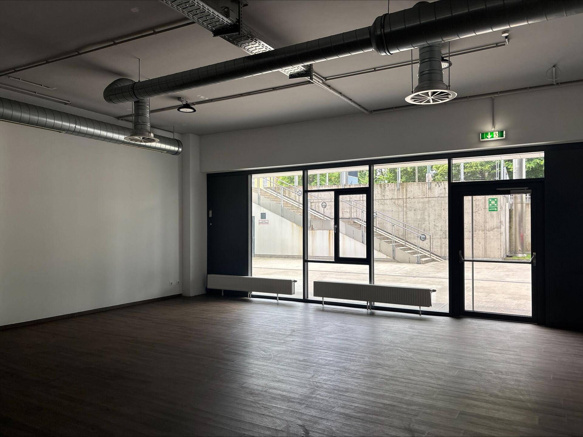 Heller, leerer Gewerberaum mit großen Fenstern und sichtbaren Lüftungsrohren an der Decke.